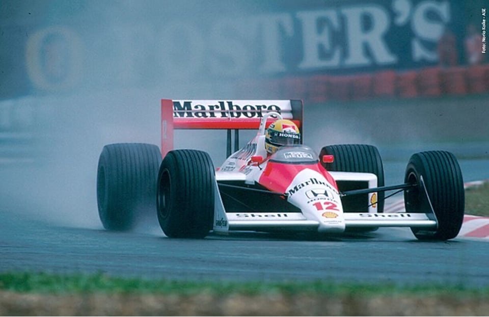 Ayrton Senna driving a red and white McLaren MP4/4 through standing water, seen from the front and slightly to the side. Spray plumes off the rear tyres and hangs in the air, blurring a Foster's advertising hoarding in the background. The car carries the number 12 and Marlboro, Honda and Shell markings, and Senna's yellow, green and blue helmet is visible in the open cockpit. A credit printed down the right-hand edge of the photograph reads Foto: Norio Koike - AGE.